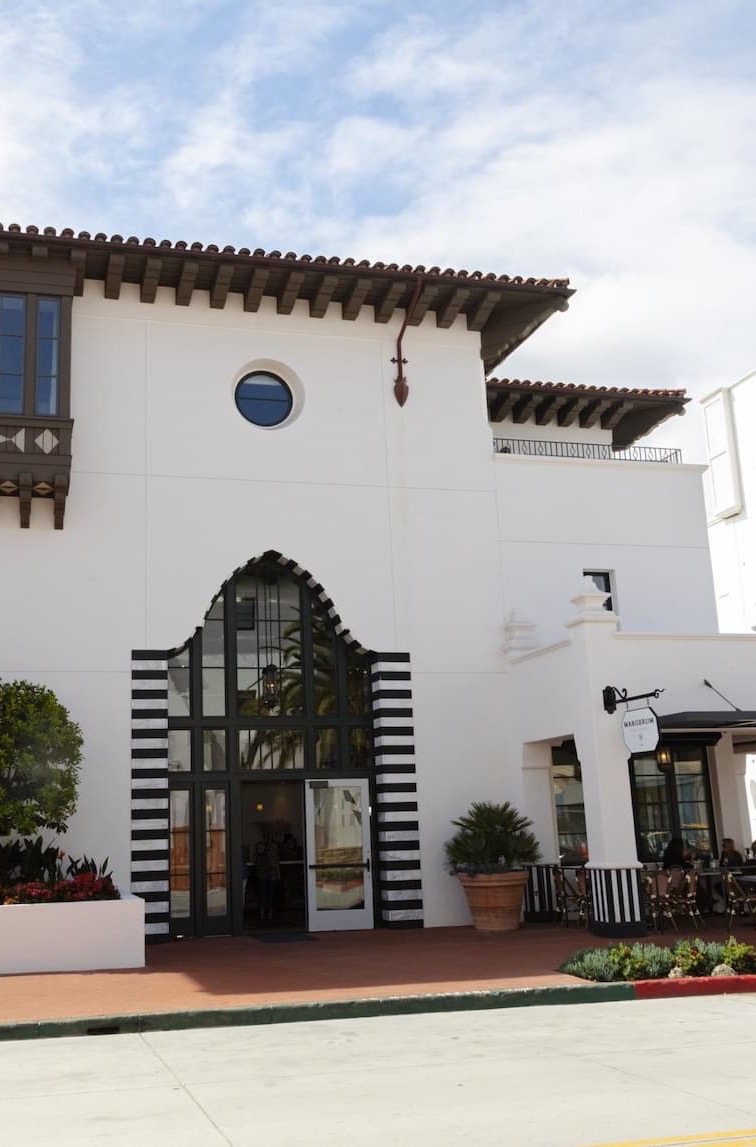 A white stucco building with Spanish and Moorish architectural details features a tall arched entry framed in black-and-white stripes, a clay tile roof, and a small outdoor café seating area along the side.