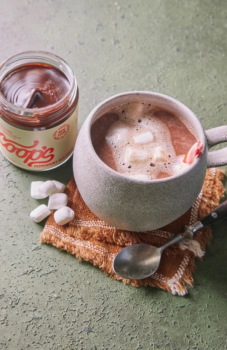 A mug of hot chocolate topped with marshmallows sits beside an open jar of peppermint chocolate sauce, with a spoon and cloth napkin on a textured green surface.
