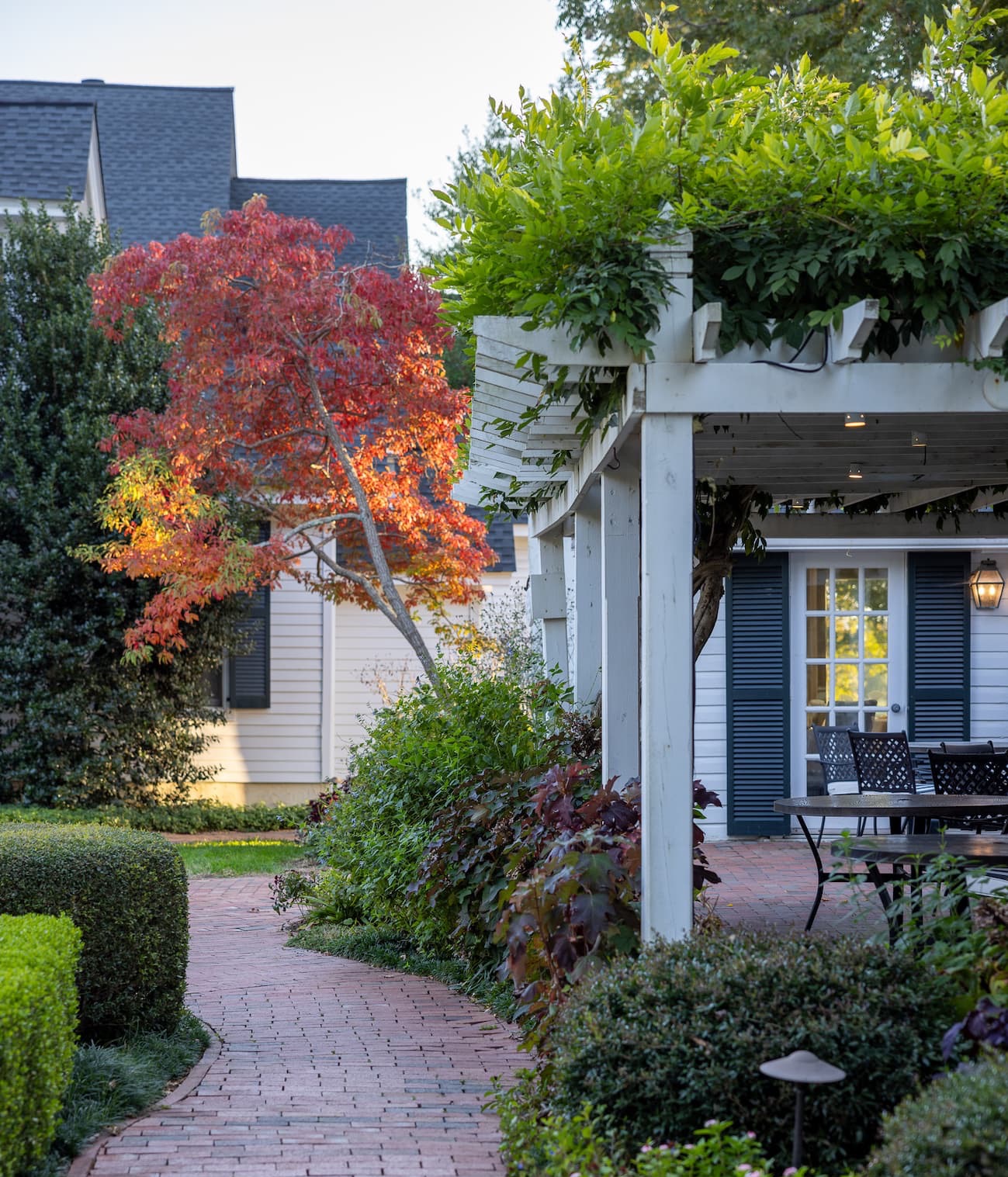 Brick pathway winding through lush gardens toward a vine-covered pergola and white cottages with vibrant autumn foliage. Fearrington Village.