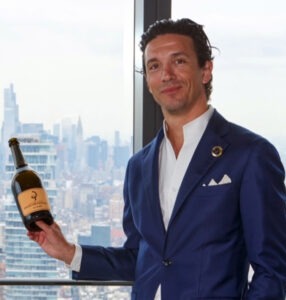 A man in a blue suit stands by a window with a city skyline in the background, smiling while holding a wine bottle with a beige label and black logo. Fearrington Village