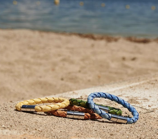 Three braided leather bracelets in yellow, brown, green, and blue rest on sunlit sand near the water, styled in a relaxed coastal setting with soft waves blurred in the background.