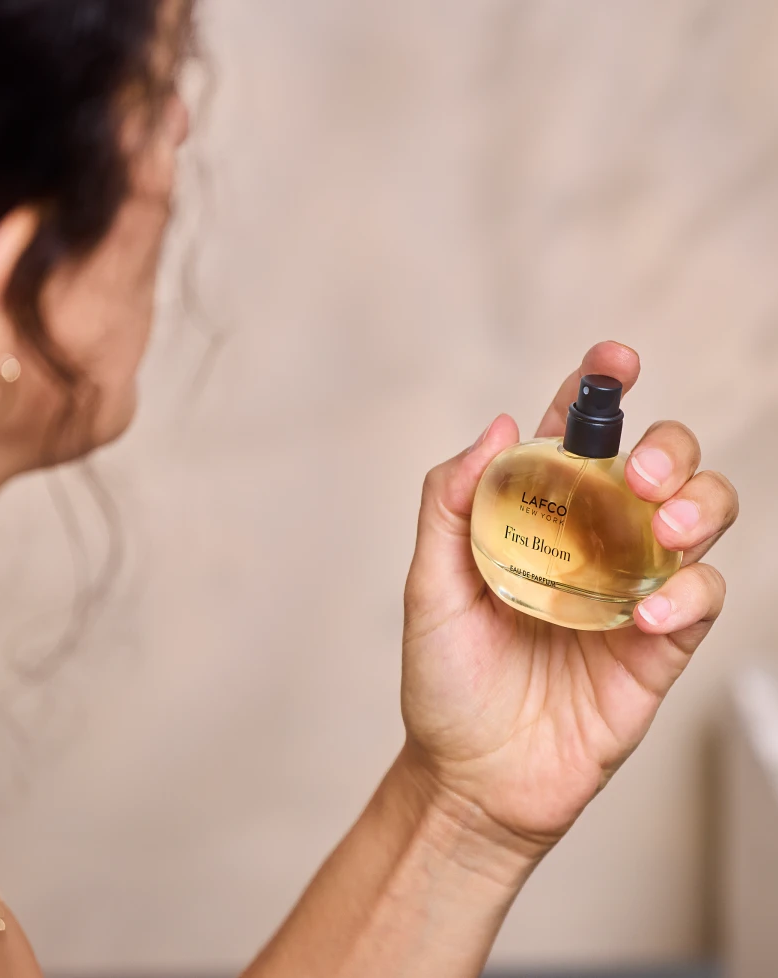 Woman holding a bottle of LAFCO First Bloom eau de parfum beside a softly glowing candle. Fearrington Village.