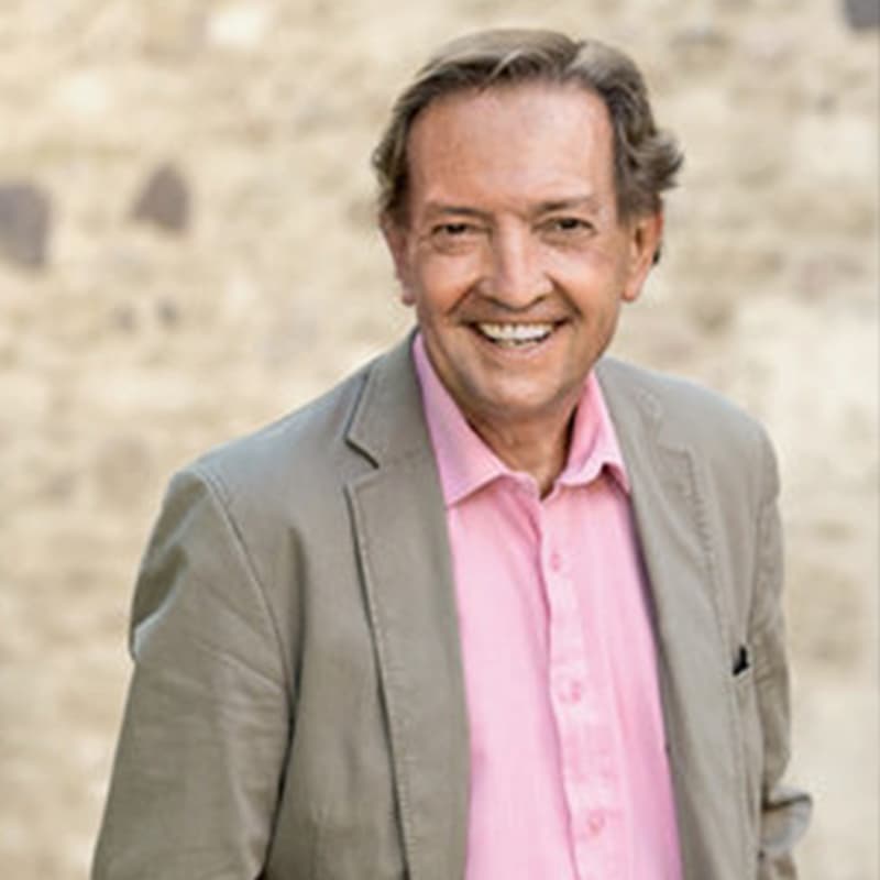 Man in a light blazer and pink button-down shirt smiles warmly at the camera. He stands against a softly blurred stone background, giving the portrait a relaxed, approachable feel.