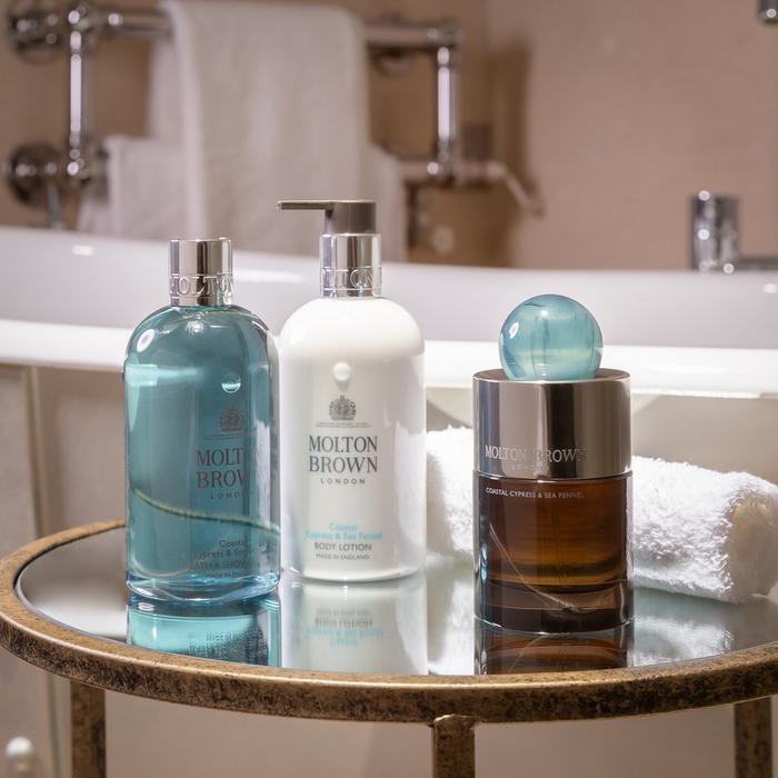 Molton Brown Coastal Cypress & Sea Fennel bath and body collection styled on a tray beside a bathtub, featuring shower gel, body lotion, and fragrance with soft towels in the background.