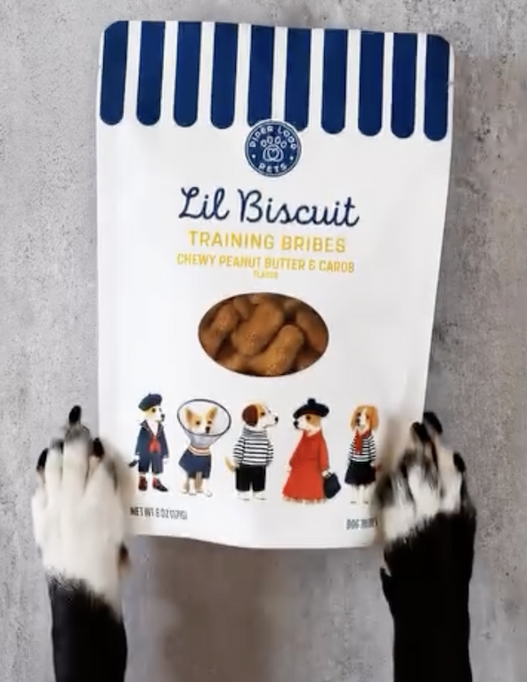Bag of Lil Biscuit Training Bribes chewy peanut butter and carob dog treats, with a dog's paws reaching toward the package on a gray surface.