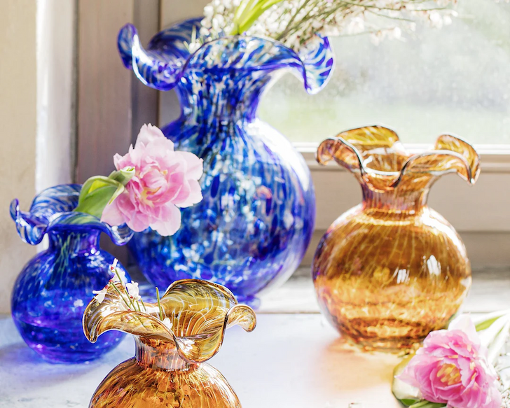 Colorful handblown glass vases in blue and amber sit by a window, styled with soft pink blooms in a bright, airy setting.