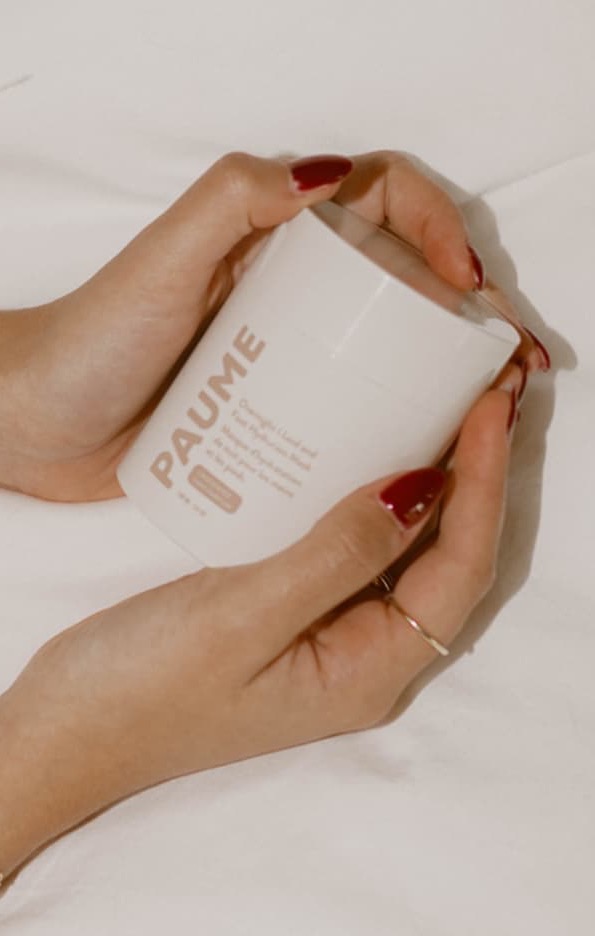 Close-up of manicured hands with red nails holding a minimal white perfume bottle, wearing delicate gold jewelry and a feather-trimmed sleeve against a soft neutral background.