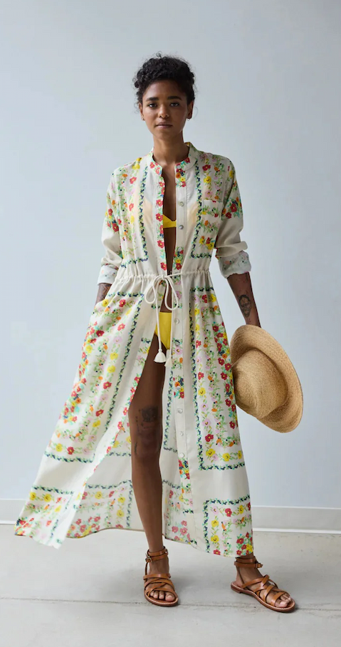 Model wearing a lightweight white floral cover-up with a drawstring waist over a yellow swimsuit, holding a woven straw hat and wearing brown leather sandals. Fearrington Village.