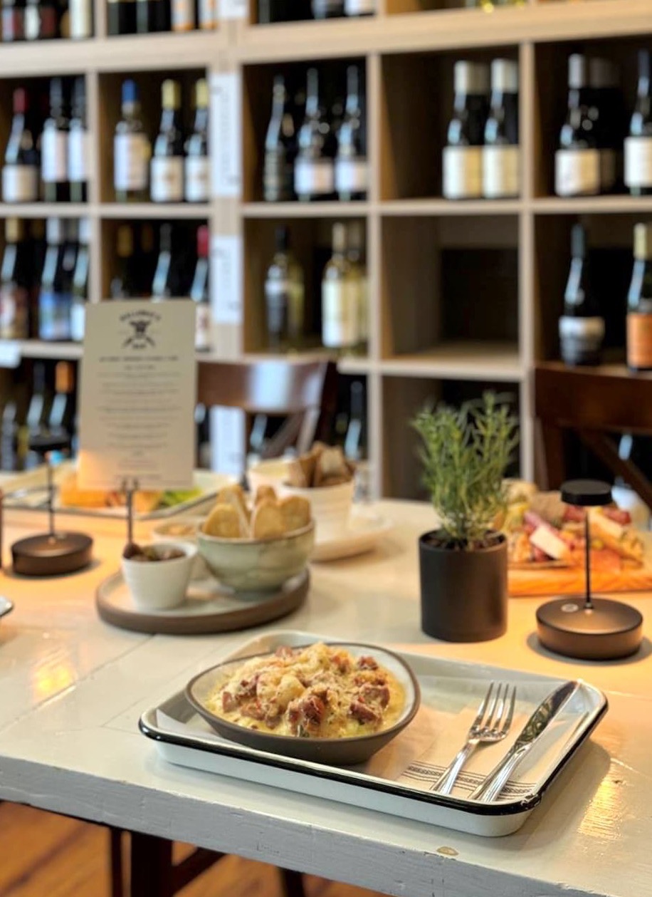 A table set with plated food, cutlery, small potted herbs, and wine glasses sits in front of shelves filled with various bottles of wine in a cozy restaurant setting. Fearrington Village