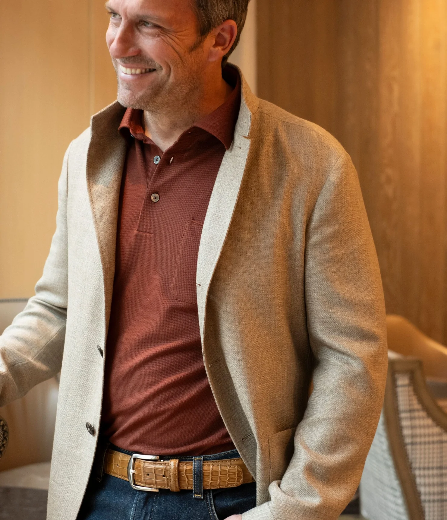 Smiling man in a beige blazer and rust polo shirt, wearing a tan textured leather belt with jeans in a warmly lit interior setting.