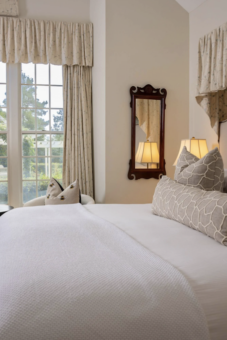 Bright, airy bedroom featuring a large bed with crisp white linens and patterned gray pillows, framed by tall windows with soft floral drapery, bedside lamps, an upholstered chair, and a decorative wood-framed mirror on a vaulted wall.