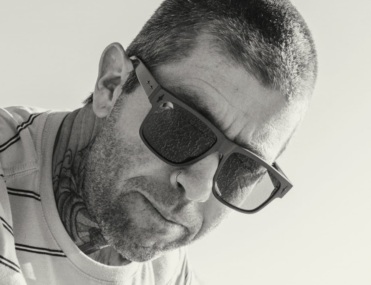 Black-and-white close-up of a man with a buzz cut and neck tattoos wearing dark sunglasses, looking downward.