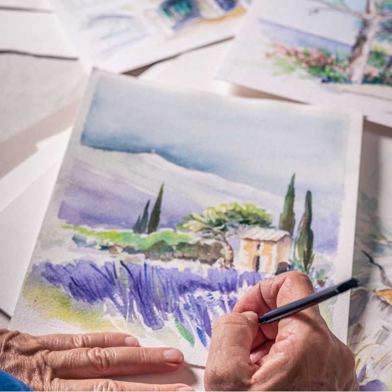 A close-up of hands painting a watercolor scene of a lavender field with a small house and tall trees, surrounded by other soft, pastel landscape sketches on a work surface.