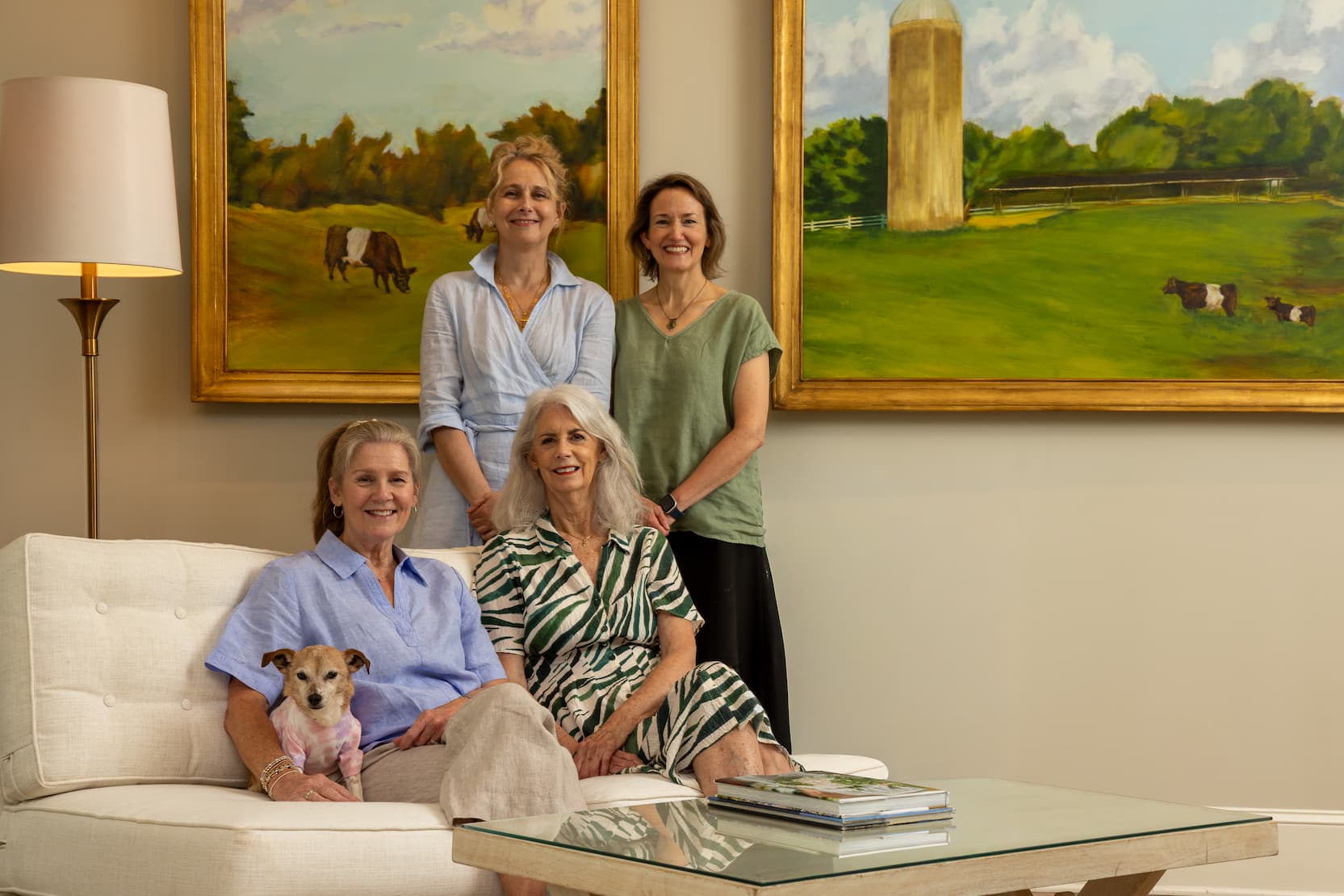 Four women gather in a bright living room with a small dog, seated and standing beneath large paintings of Fearrington's Belted Galloway cows and iconic silo, reflecting the warm hospitality and community spirit of the village. Fearrington Village.