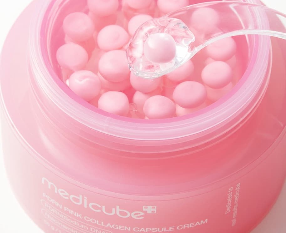 Open jar of Medicube pink collagen capsule cream with a spatula lifting a single gel capsule from the container.
