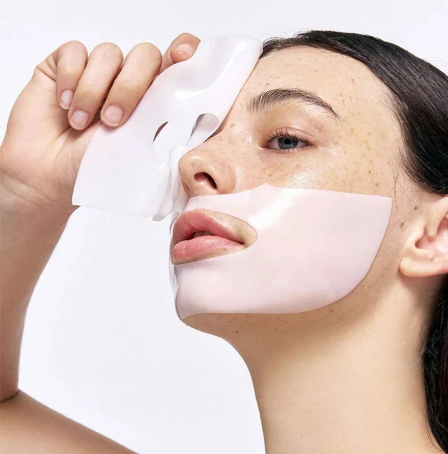 Close-up of a person peeling a pale pink sheet mask upward from their forehead while the rest of the mask remains on their face.