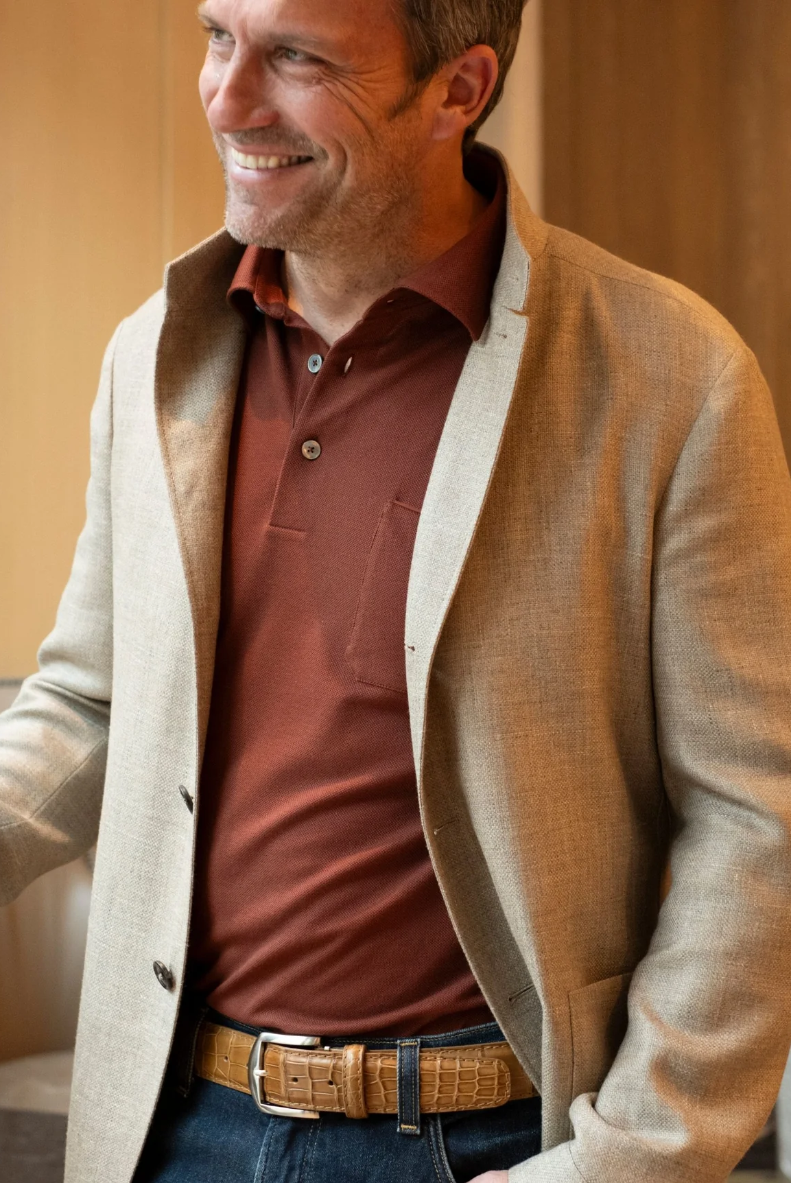 Smiling man wearing a rust polo shirt, light tan blazer, and textured leather belt, standing indoors in warm natural light.