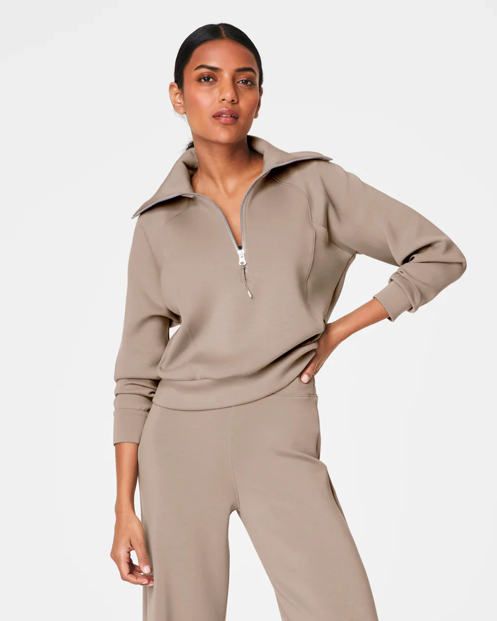A person with short hair is wearing a SPANX AIRESSENTIALS HALF ZIP sweater in navy blue, made from lightweight spacer fabric, paired with black pants. Their arms are crossed over their chest as they stand against a plain, light grey background. Fearrington Village
