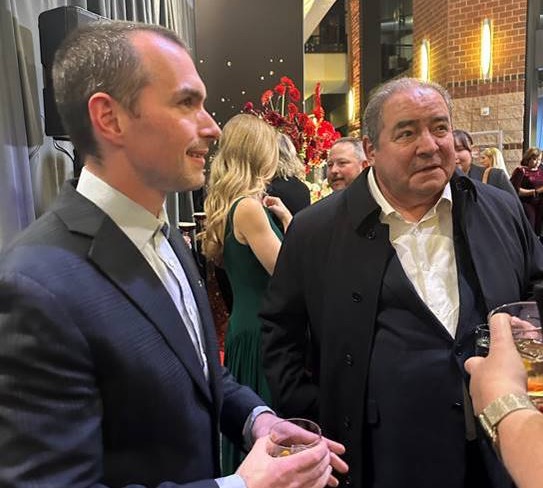Two men in suits stand indoors at a social event, holding drinks and conversing. There are other people in formal attire in the background, with red floral decorations and warm lighting. Fearrington Village