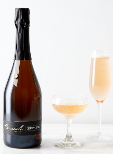 Bottle of Caraccioli Brut Rosé sparkling wine beside a coupe and a flute, each filled with pale pink bubbly, set on a light tabletop against a neutral background.