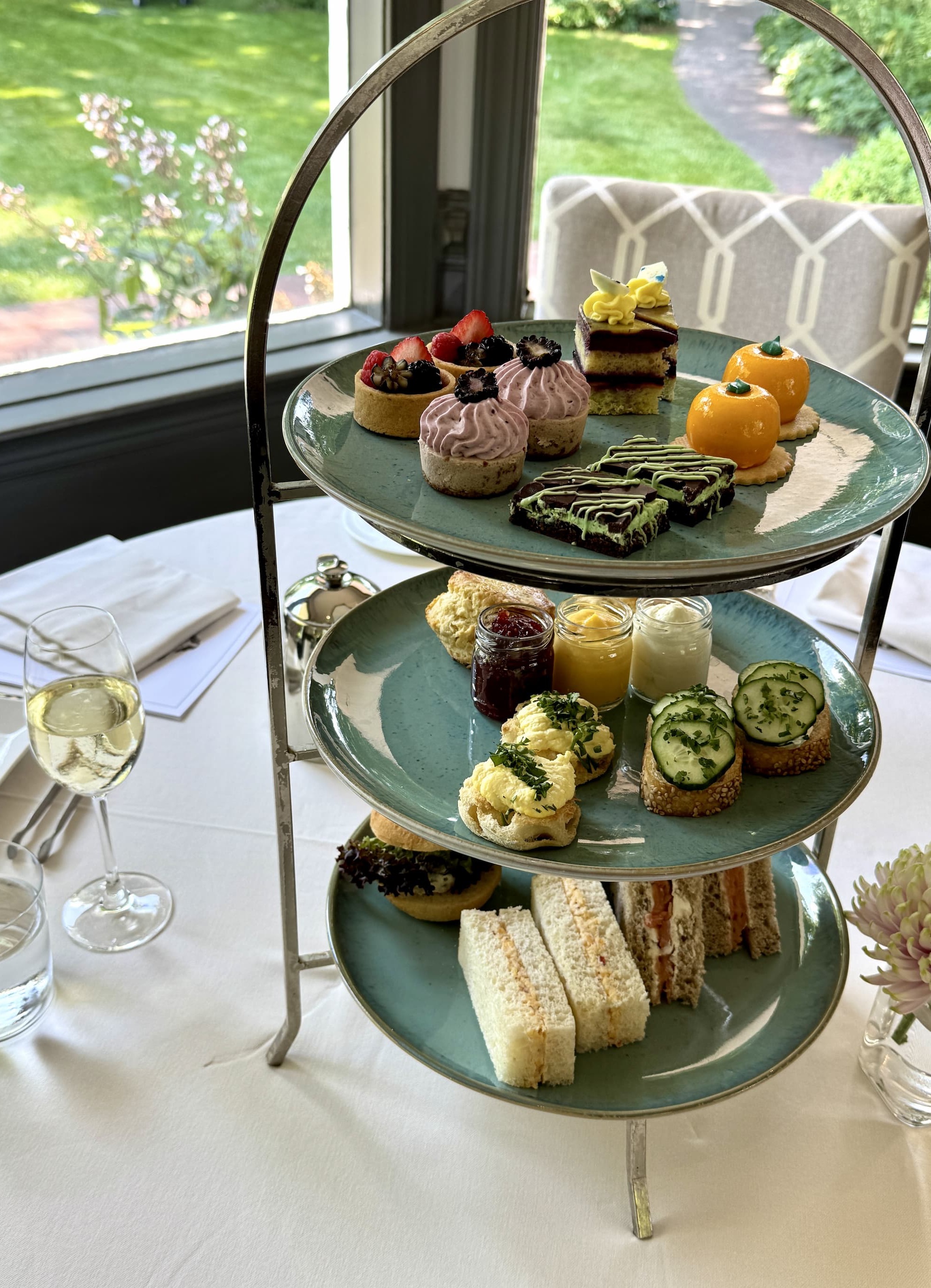 Three-tier afternoon tea stand filled with tea sandwiches, pastries, fruit tarts, cakes, and savory bites, set on a white tablecloth beside a glass of sparkling wine with garden views through large windows.