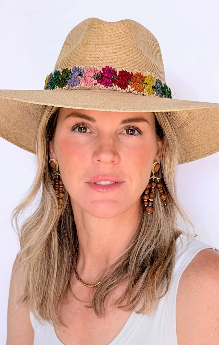Woman wearing a natural straw hat with a hand-embroidered multicolor floral band, paired with beaded drop earrings and a white sleeveless top, against a clean white background. Fearrington Village.