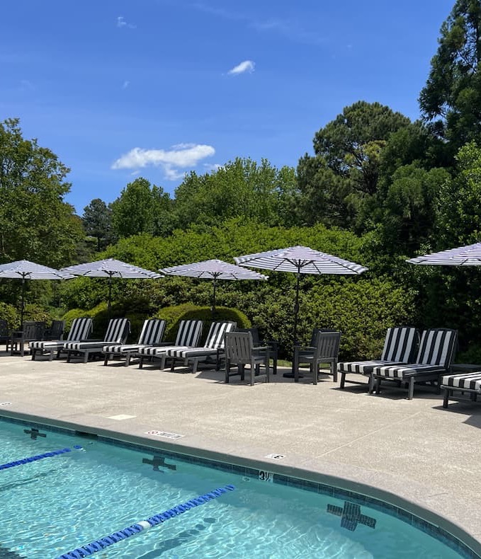 A sunny outdoor swimming pool is lined with black-and-white striped lounge chairs and matching umbrellas, creating a classic resort-style atmosphere. Surrounded by manicured shrubs and mature trees, the pool deck offers a peaceful setting for relaxation. Clear blue water, bright skies, and lush greenery contribute to the inviting, summer-ready scene.