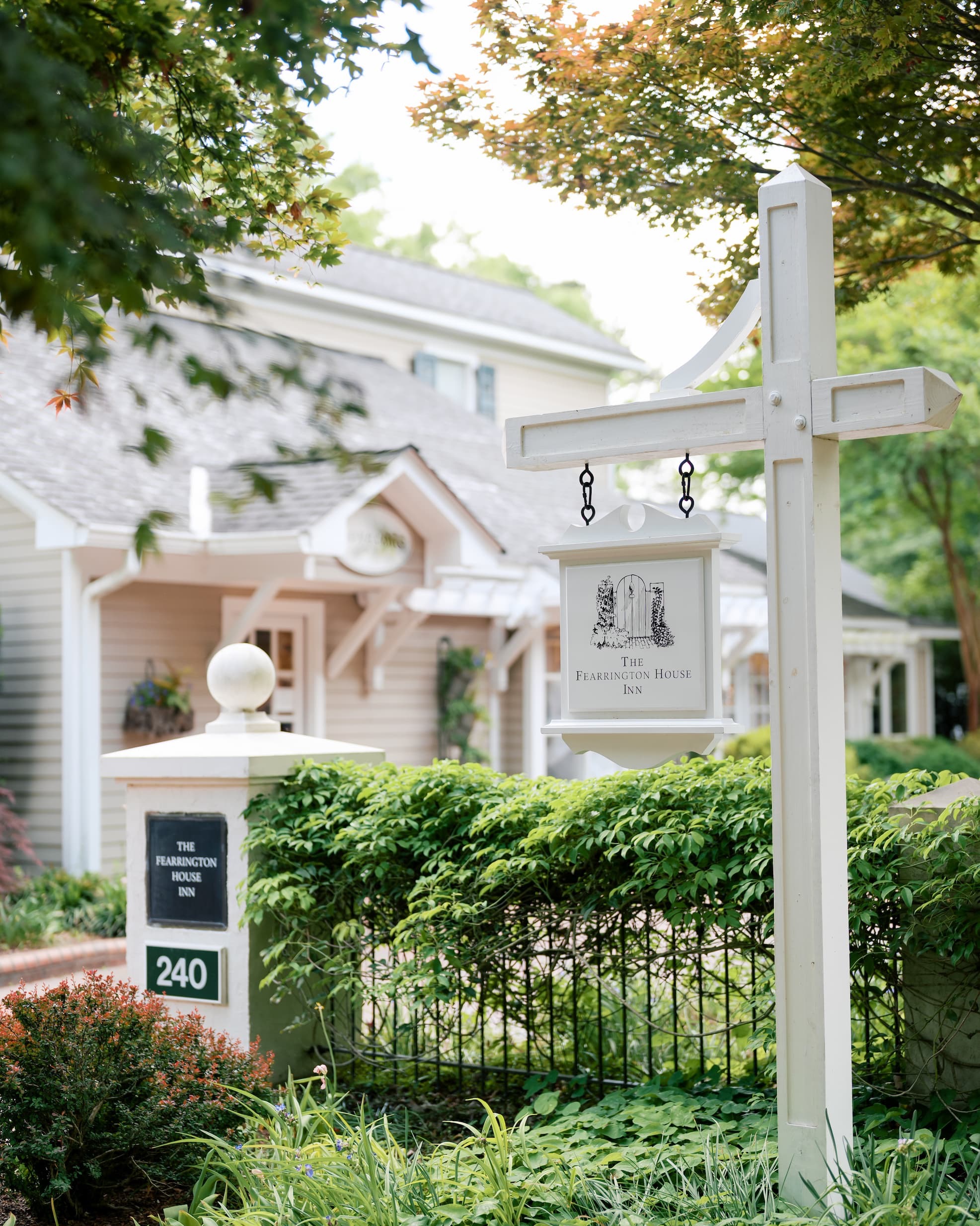 White sign for The Fearrington House Inn standing among lush gardens and manicured hedges, with the inn’s charming cottage-style buildings softly blurred in the background beneath leafy trees.
