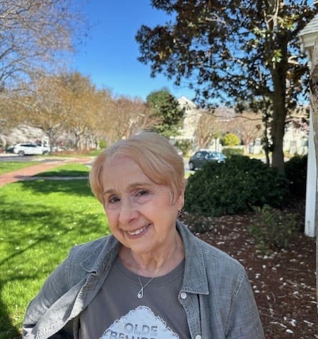 An older woman smiles outdoors on a sunny day, standing near a grassy area with trees and buildings in the background.