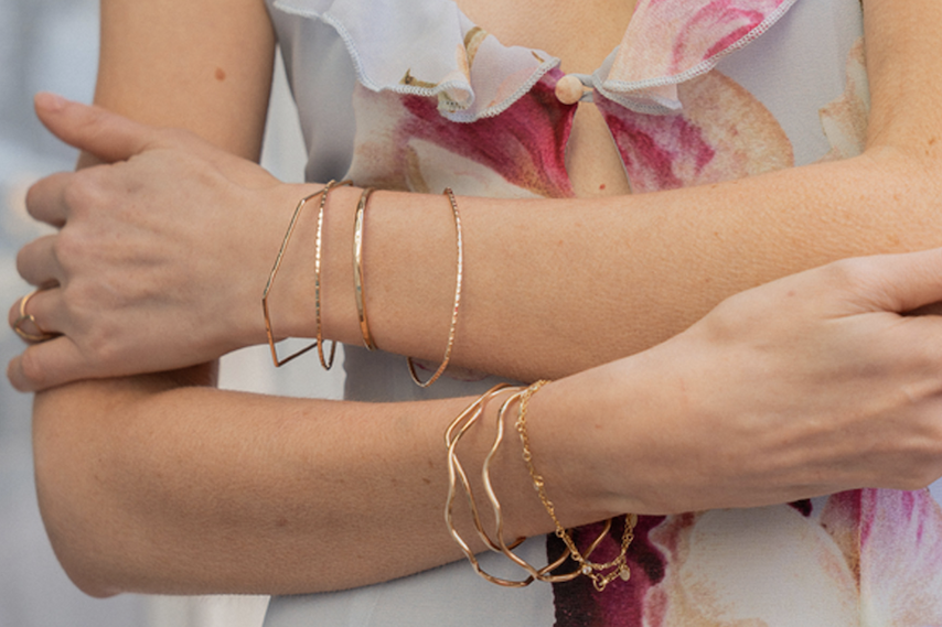 Close-up of crossed arms wearing multiple delicate gold bracelets—thin bangles, a geometric cuff, and chain styles—paired with a light, floral-patterned blouse.