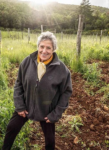 Smiling woman standing in a cultivated field with rows of plants and wooden posts, with wooded hills in the background.