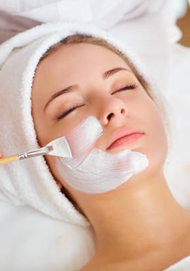 Close-up of a woman with eyes closed receiving a facial treatment, a white cream mask being applied to her cheek with a brush.