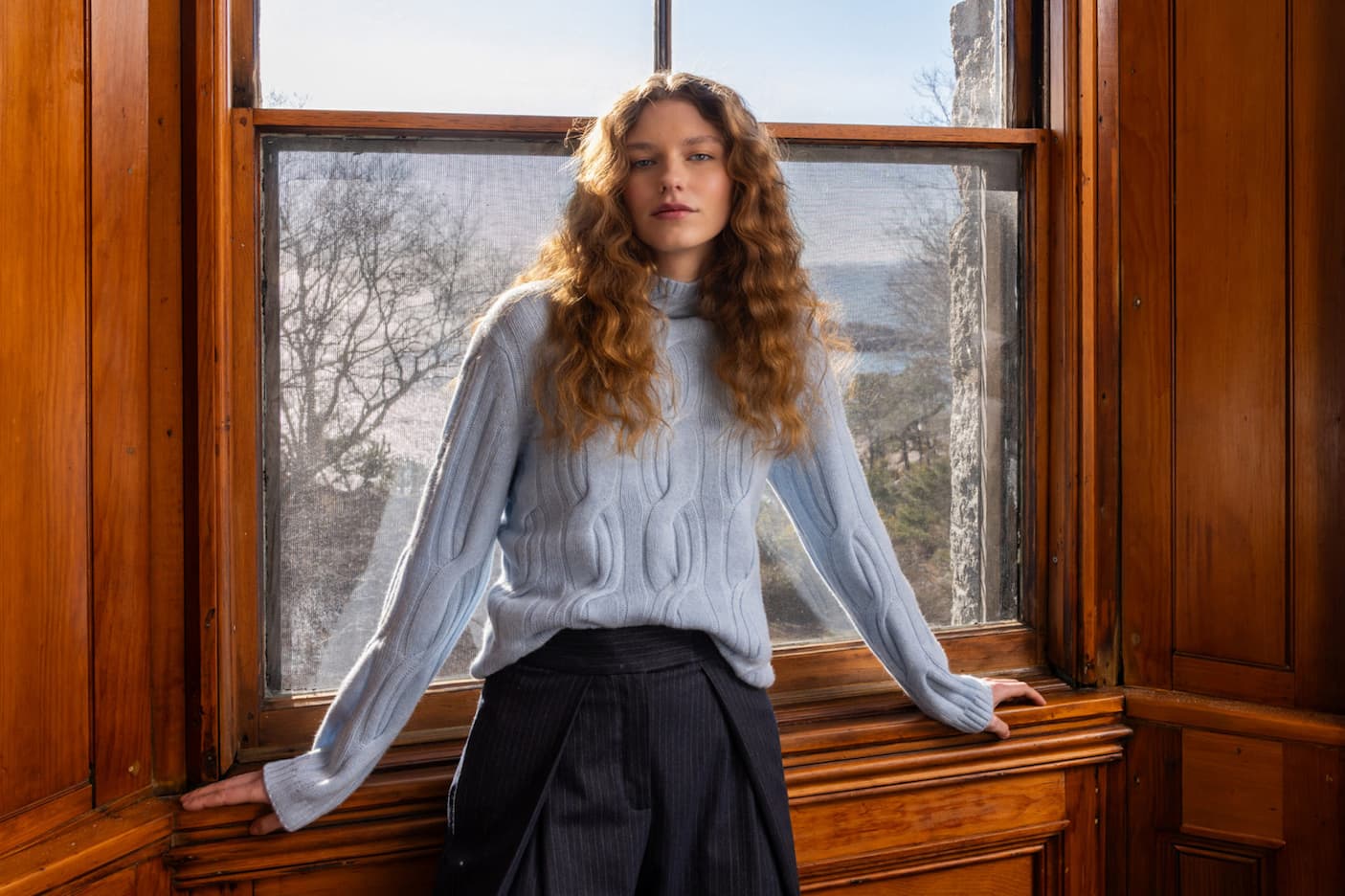 Woman wearing a light blue cable-knit sweater with dark trousers by a sunlit window. Fearrington Village.