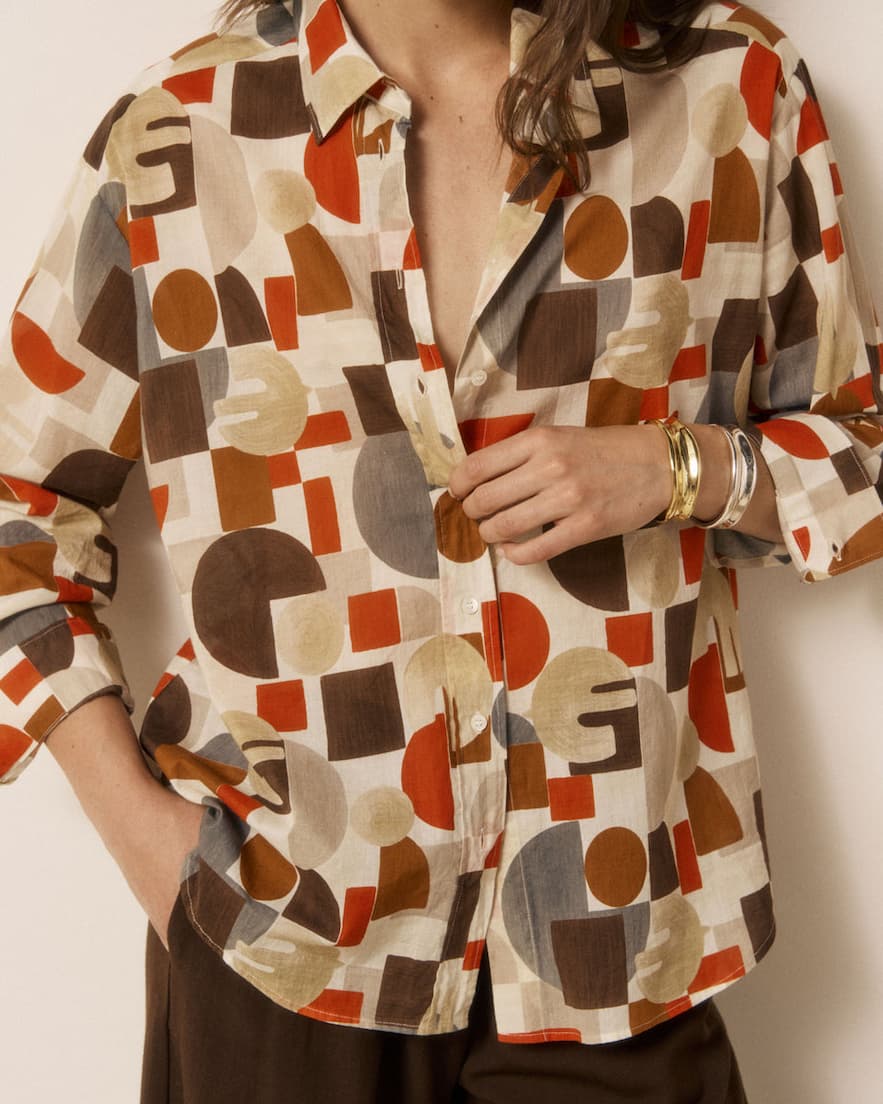 Woman wearing a geometric-print blouse in warm rust, brown, cream and gray tones with stacked bracelets. Fearrington Village.
