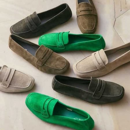 Suede penny loafers in green, taupe, beige and charcoal arranged on a neutral background. Fearrington Village.