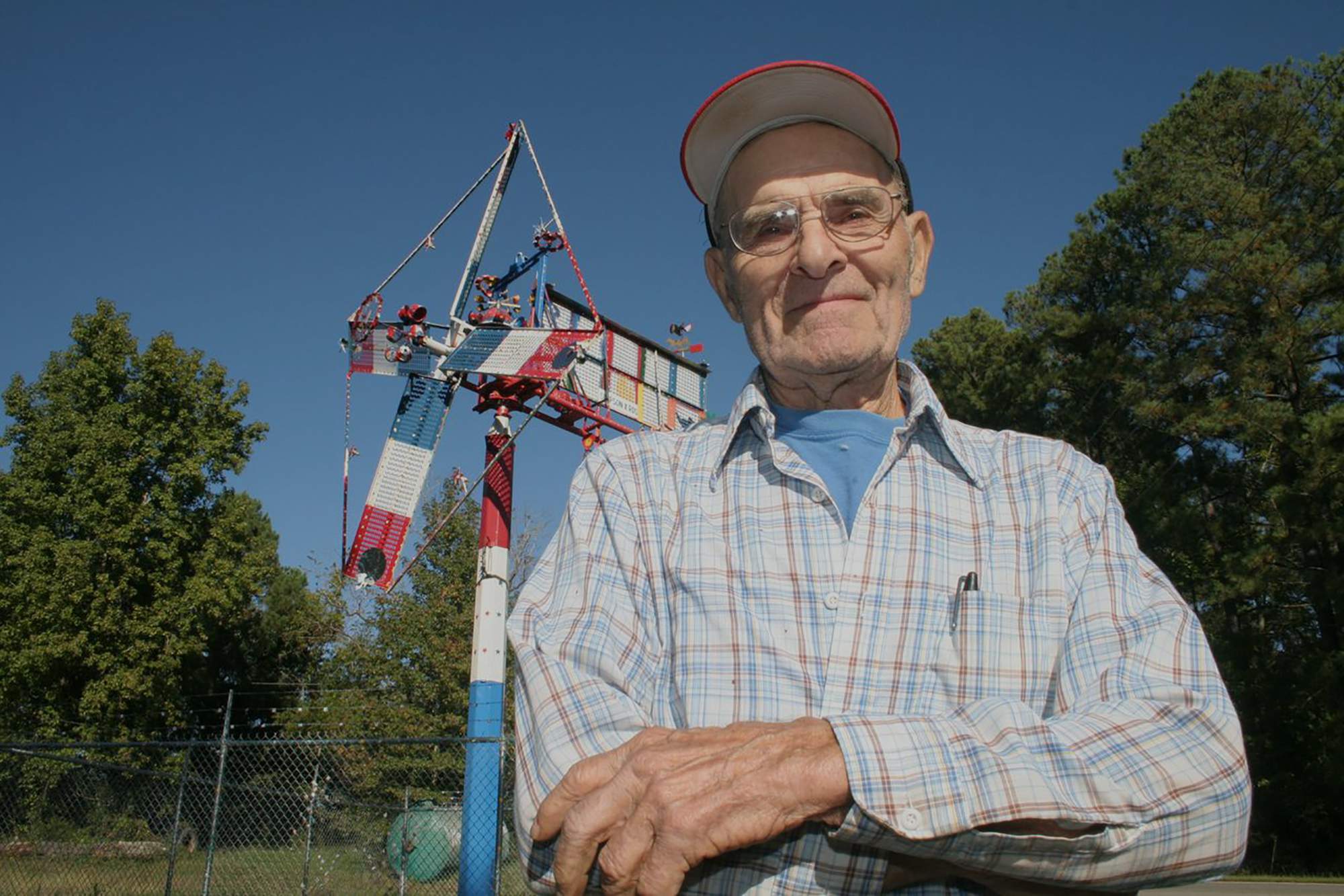 Vollis Simpson standing outdoors with arms crossed, with one of his large whirligig sculptures behind him.