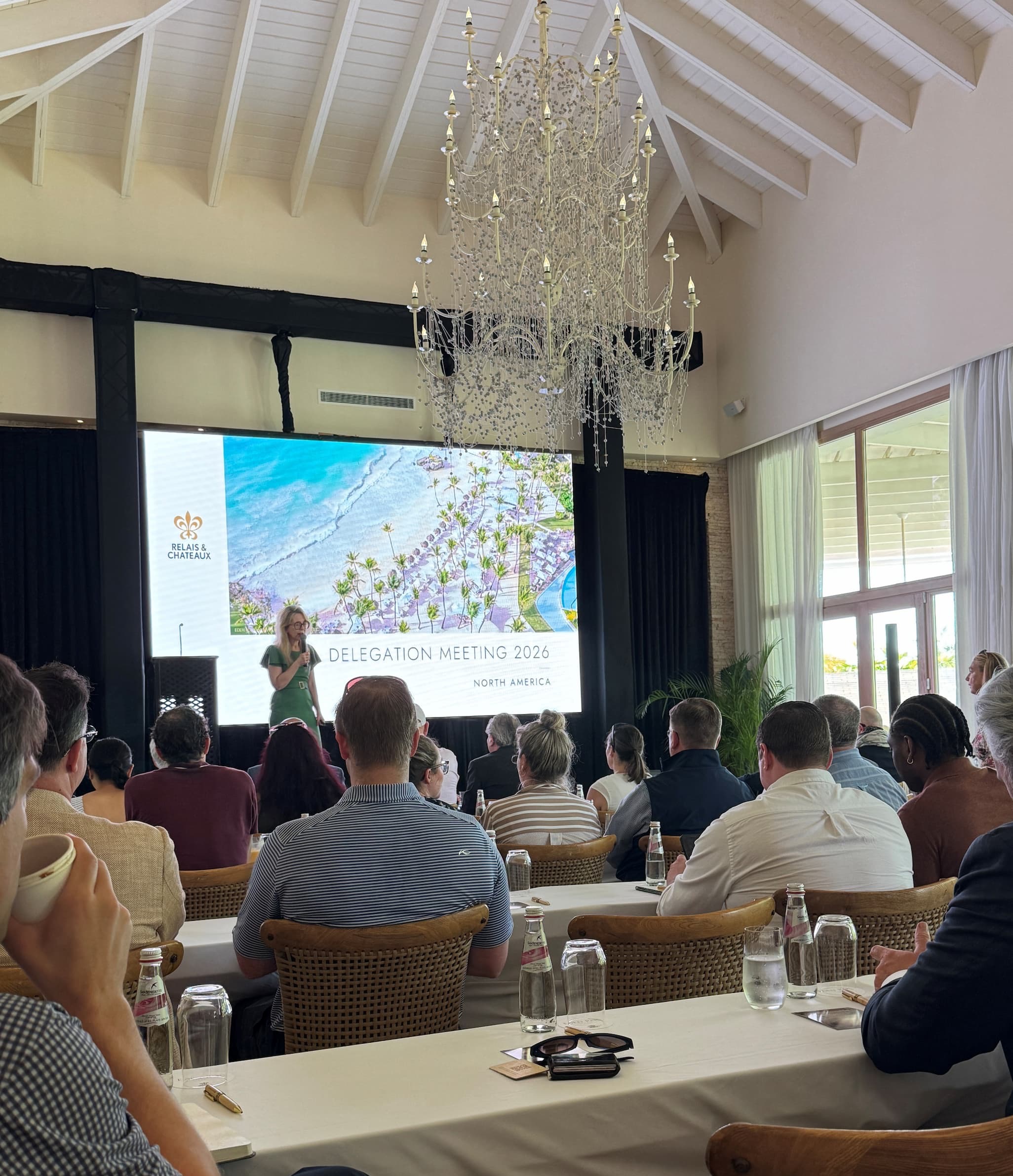 A conference-style gathering in a bright, high-ceilinged room with exposed beams and a large chandelier. Attendees sit at long tables facing a stage where a speaker presents in front of a large screen displaying “Relais & Châteaux Delegation Meeting 2026 – North America” alongside an aerial coastal image. Natural light pours in from tall windows on the right, and water bottles, glasses, and notepads are arranged neatly on the tables.