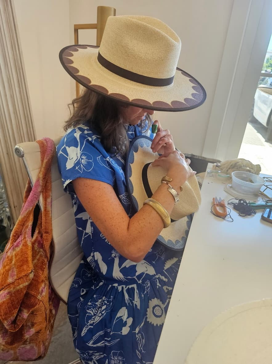 A woman in a blue floral dress hand-stitches the brim of a wide-brim straw hat with blue scalloped trim while seated at a worktable filled with crafting supplies in a bright studio. Fearrington Village.