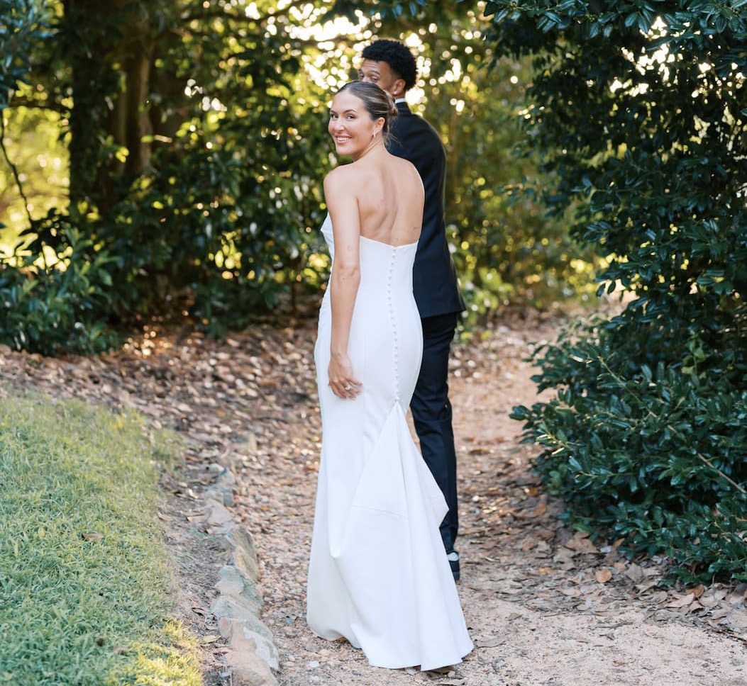 Bride in a strapless white gown smiles back over her shoulder while walking along a garden path, with the groom in a dark suit just behind her among leafy greenery.