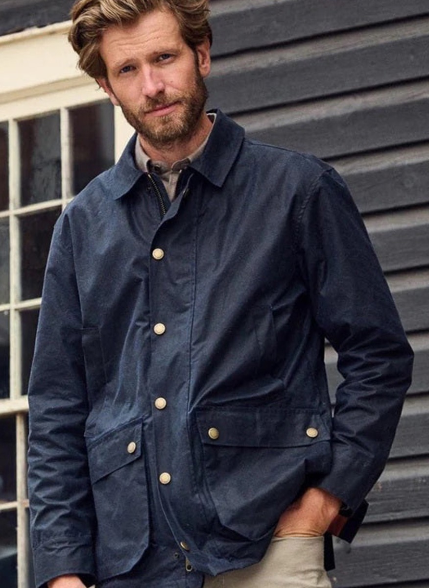 A man with light brown hair and a beard is standing in front of a dark wooden building, wearing a navy blue jacket with large buttons and beige pants. One hand is in his pocket and he is looking at the camera. Fearrington Village