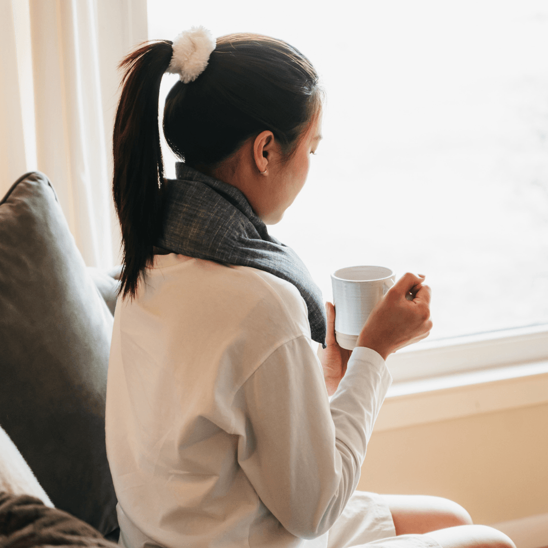 A person with their hair in a ponytail sits by a bright window, holding a white mug and looking outside, creating a quiet, reflective morning-at-home moment