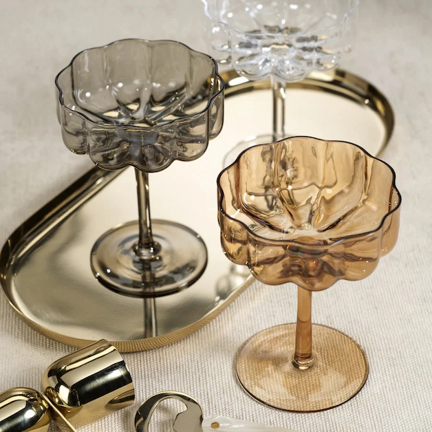 Amber, smoky gray and clear scalloped coupe glasses arranged on a reflective gold serving tray with cocktail accessories. Fearrington Village.