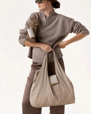A woman in a monochromatic taupe outfit carries an oversized beige tote bag with a matching attached pouch. The lightweight bag drapes softly from her shoulder, complementing a relaxed knit sweater, cargo-style pants, sunglasses, and gold accessories for a modern, understated look against a neutral background.