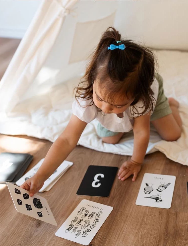 A young child with a blue hair bow sits on the floor, reaching for a black-and-white flashcard among several cards spread out, including one with the number three and others with images and sign language. Fearrington Village