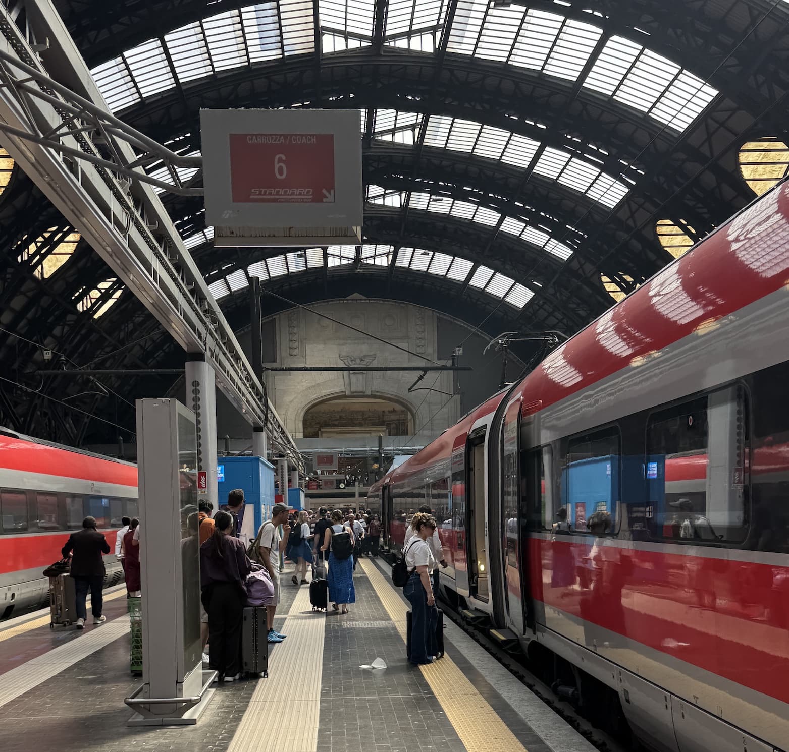Travelers wait beside a red high-speed train at a busy station platform beneath a soaring glass-and-steel arched roof. A sign overhead marks Coach 6 as passengers board with suitcases and backpacks.