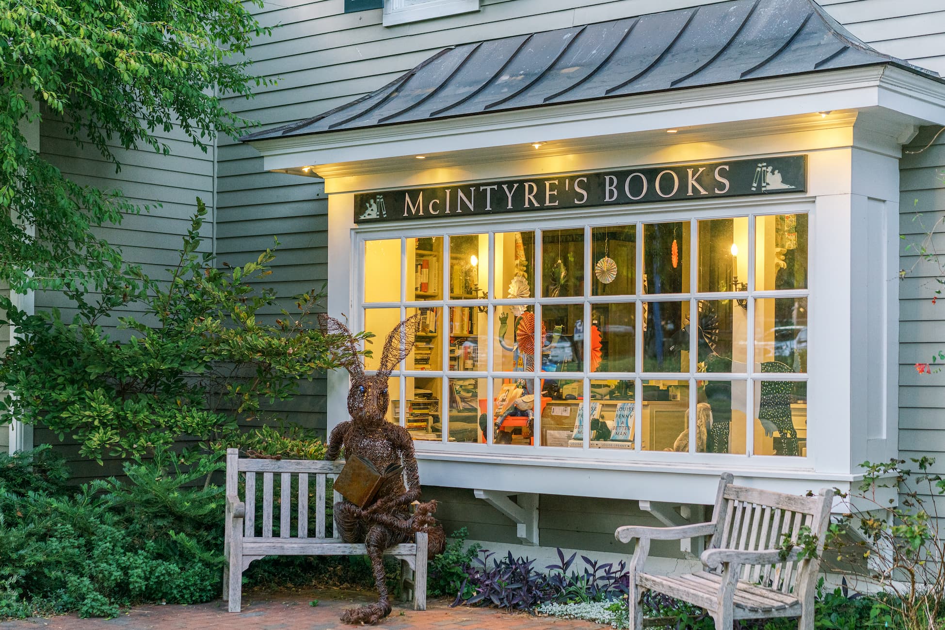 Exterior of McIntyre’s Books with warmly lit windows, a wooden bench, and a whimsical rabbit sculpture reading a book in a garden setting.