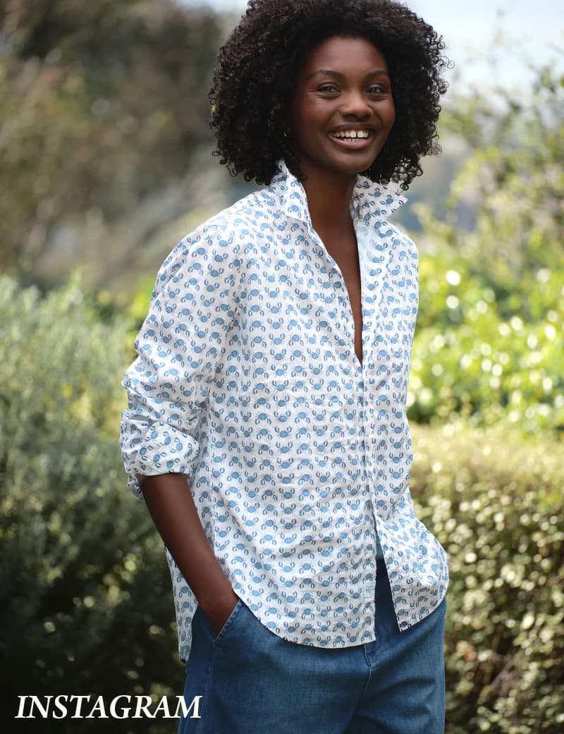 Smiling woman with natural curly hair wearing a light patterned button-down shirt and jeans, standing outdoors with hands in her pockets, set against softly blurred garden greenery.