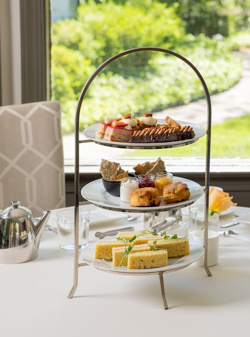 Elegant three-tier afternoon tea stand featuring finger sandwiches, fresh scones with preserves and cream, and an assortment of pastries and desserts, set on a white tablecloth beside a silver teapot and garden-view windows.