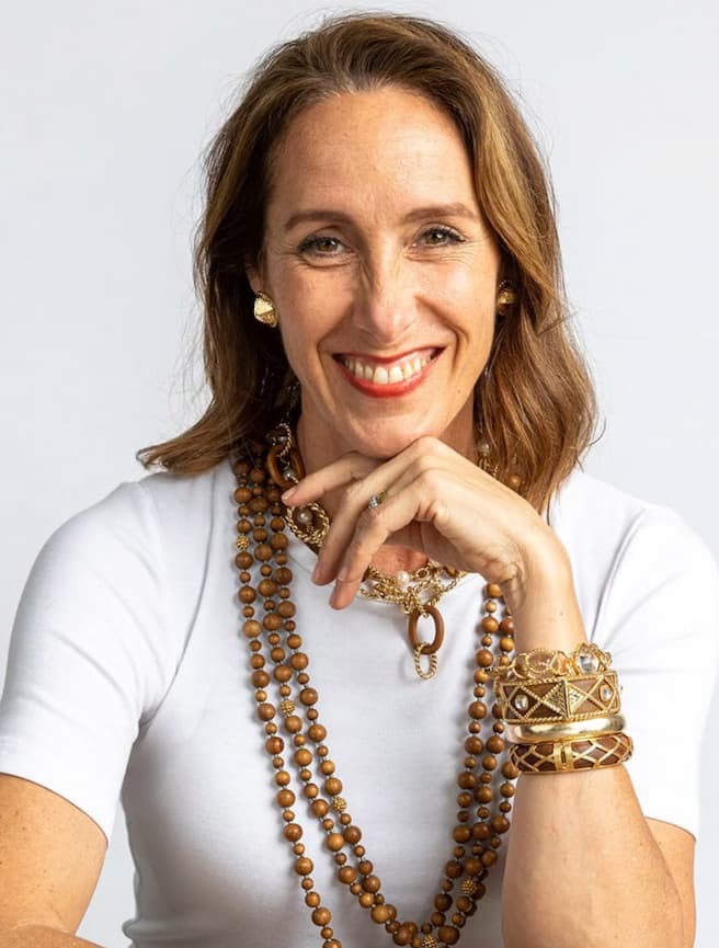 Smiling woman with shoulder-length brown hair wearing a white top and layered gold and wooden bead necklaces, stacked gold bracelets, and gold earrings, resting her chin on her hand against a plain light background.
