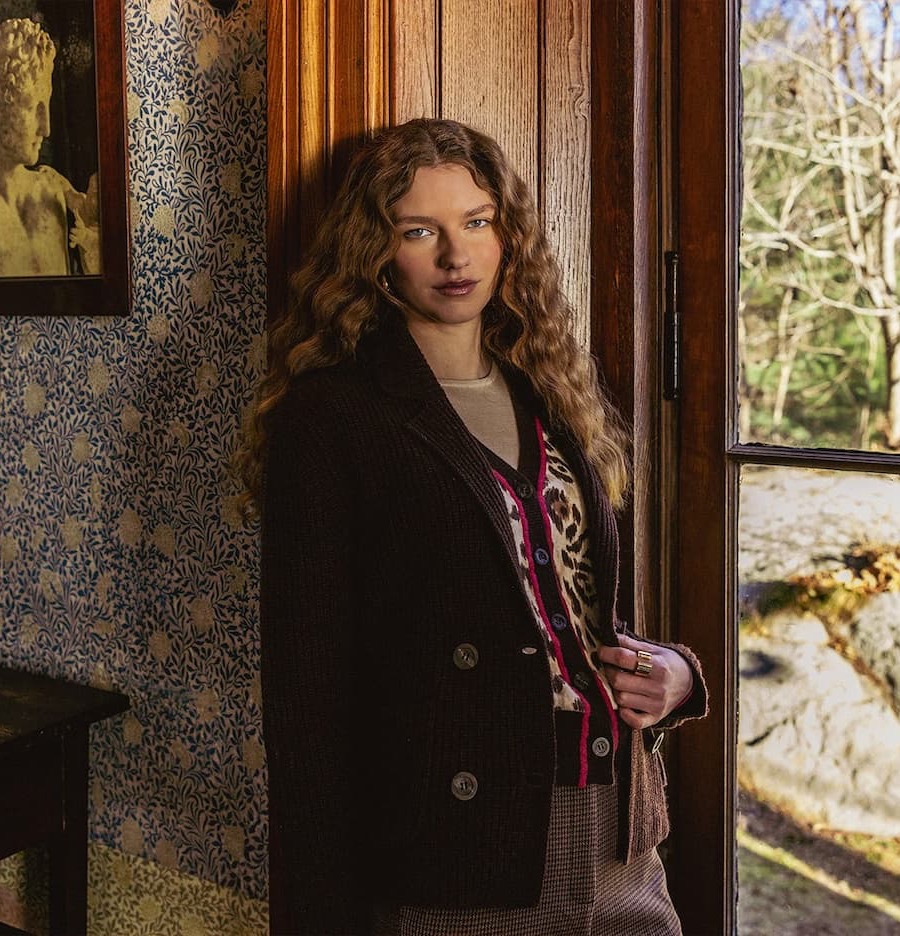 A woman models layered fall fashion beside a sunlit window in a richly decorated interior. Fearrington Village.